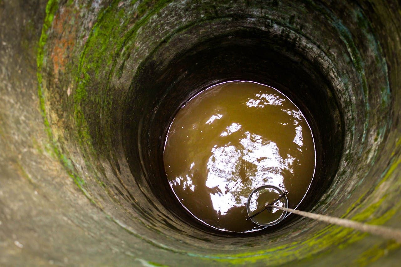 Почему тампонаж обязателен при ликвидации скважин A view down an old stone well showing murky water and a rope with a bucket.